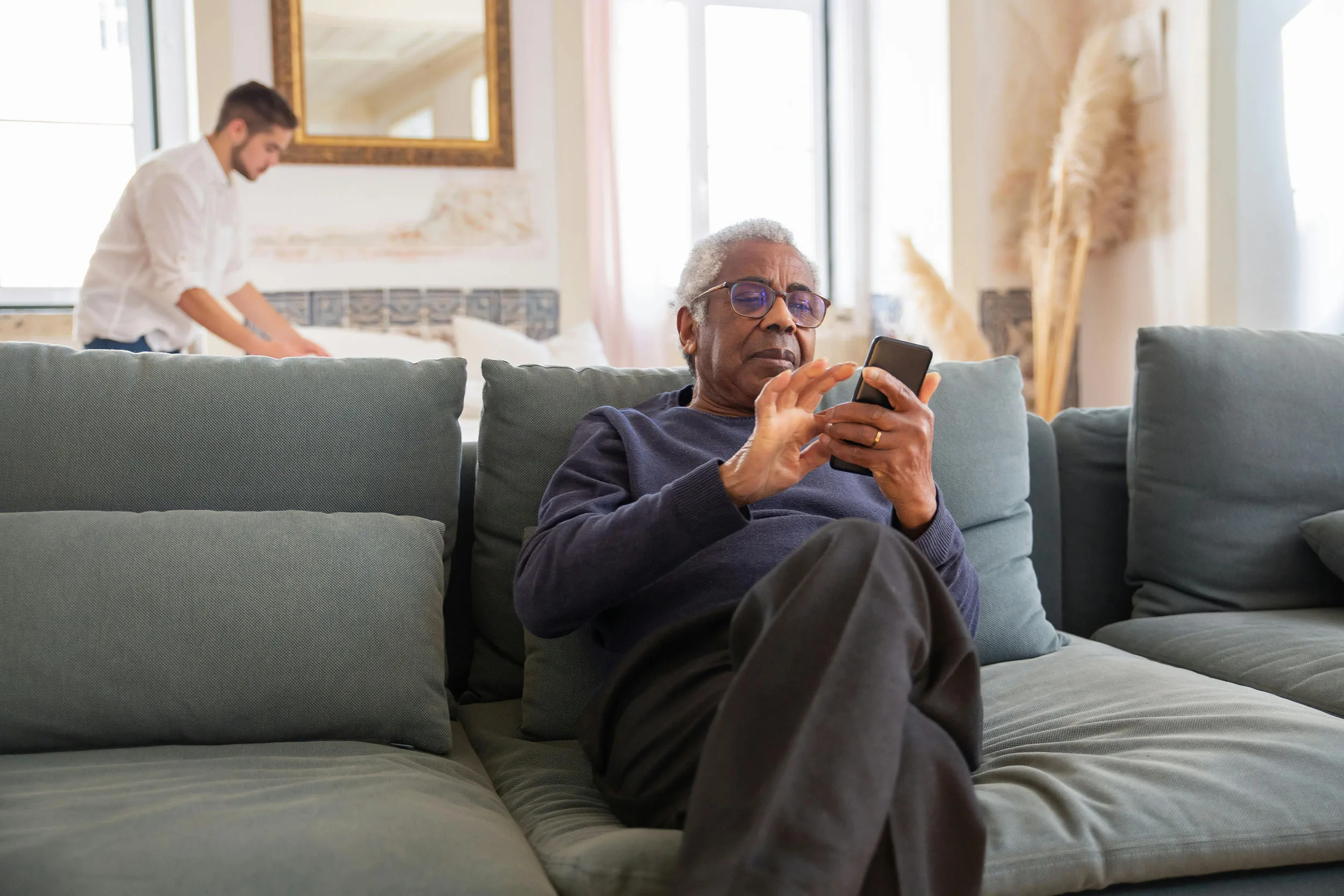 Senior man using a smartphone while a caregiver arranges a bed in the background.